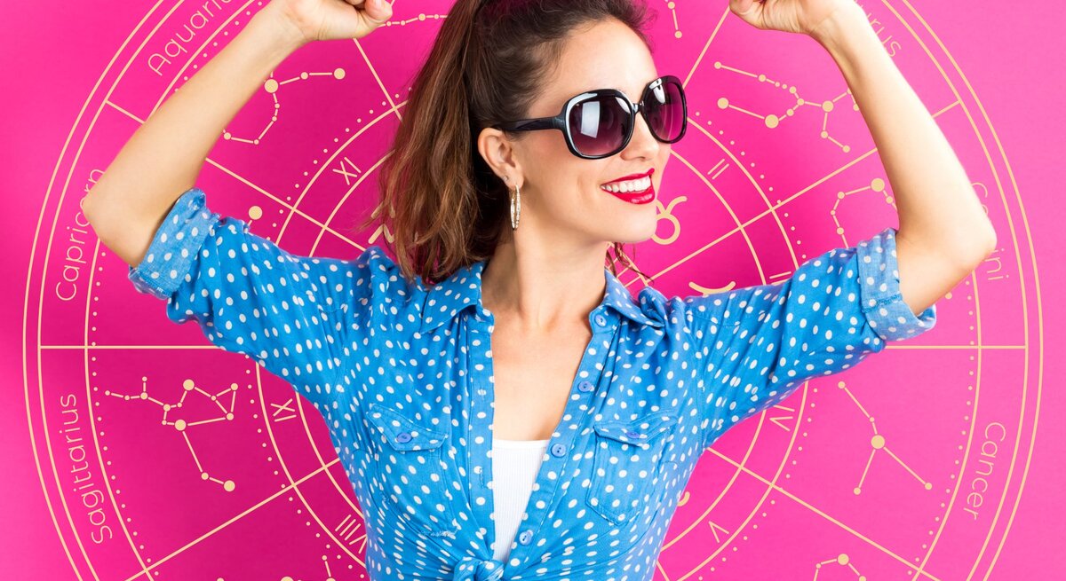 Eine Frau in einer blauen, gepunkteten Bluse posiert selbstbewusst vor einem astrologischen Rad, das verschiedene Sternzeichen zeigt. Sie trägt eine Sonnenbrille und hebt ihre Arme in einer kraftvollen Pose. Das Bild vermittelt Optimismus, Selbstbewusstsein und Erfolg.