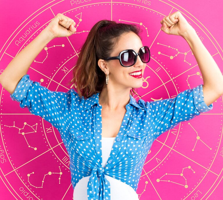 Eine Frau in einer blauen, gepunkteten Bluse posiert selbstbewusst vor einem astrologischen Rad, das verschiedene Sternzeichen zeigt. Sie trägt eine Sonnenbrille und hebt ihre Arme in einer kraftvollen Pose. Das Bild vermittelt Optimismus, Selbstbewusstsein und Erfolg. | © Getty Images/ Melpomenem und Chikovnaya via Canva.com [M]