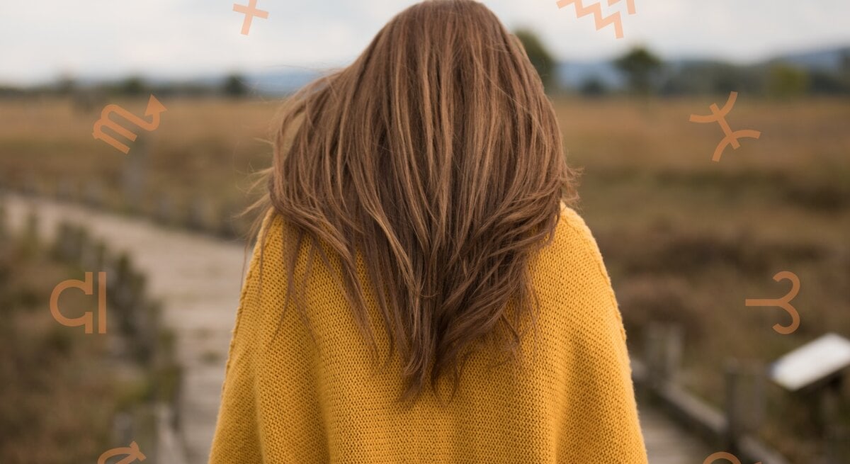 Eine Frau mit langen braunen Haaren in einem gelben Pullover steht auf einem Holzsteg in einer weiten Landschaft. Um ihren Kopf herum sind astrologische Symbole verschiedener Sternzeichen angeordnet.