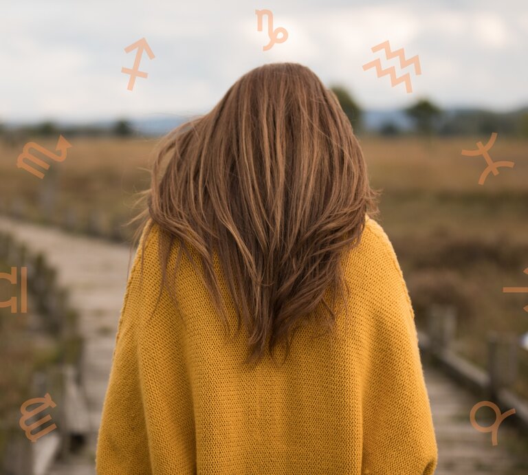 Eine Frau mit langen braunen Haaren in einem gelben Pullover steht auf einem Holzsteg in einer weiten Landschaft. Um ihren Kopf herum sind astrologische Symbole verschiedener Sternzeichen angeordnet. | © Adobe Stock/ Christian Schwier und Chikovnaya via Canva.com [M]