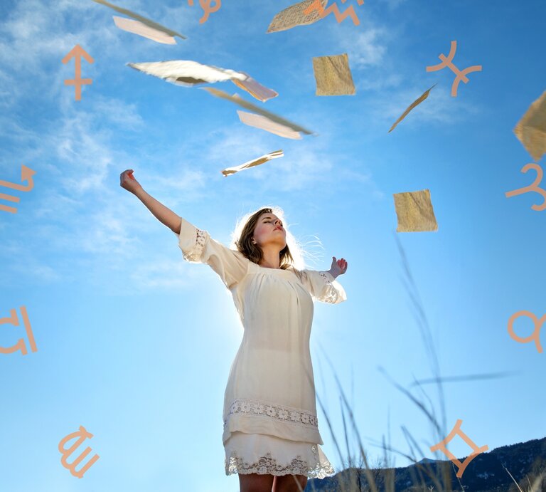 Eine Frau in einem weißen Kleid steht in einer weiten Landschaft unter einem klaren blauen Himmel. Sie hebt ihre Arme in die Luft, während um sie herum Papierblätter und Symbole der Sternzeichen schweben. Die Szene vermittelt ein Gefühl von Freiheit, Leichtigkeit und spiritueller Verbindung. | © Getty Images Signature/ DanielBendjy und Chikovnaya via Canva.com [M]
