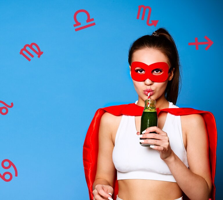 ine junge Frau trägt eine rote Superheldenmaske und ein passendes rotes Cape, während sie einen grünen Smoothie aus einer Flasche mit einem Strohhalm trinkt. Sie trägt ein weißes Sport-Top und steht vor einem blauen Hintergrund, umgeben von roten astrologischen Symbolen verschiedener Sternzeichen. | © Chikovnaya und dashapetrenkophotos via Canva.com [M]