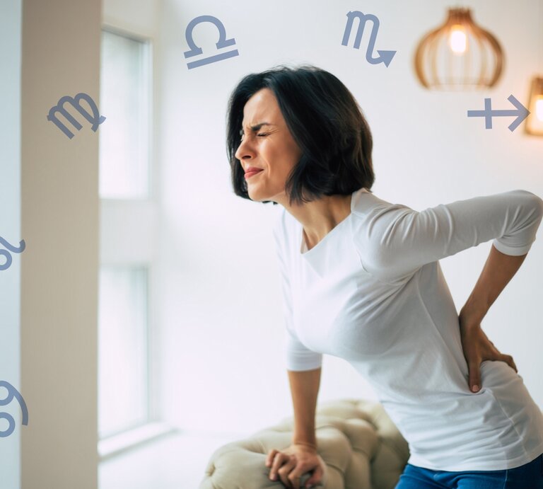 Eine Frau mit kurzen dunklen Haaren hält sich mit schmerzverzerrtem Gesicht den unteren Rücken, während sie sich an einer Couch abstützt. Um sie herum schweben verschiedene astrologische Symbole der Sternzeichen, was auf einen Zusammenhang zwischen Rückenschmerzen und den Sternzeichen hinweist. | © Getty Images/ Povozniuk und Chikovnaya via Canva.com [M]
