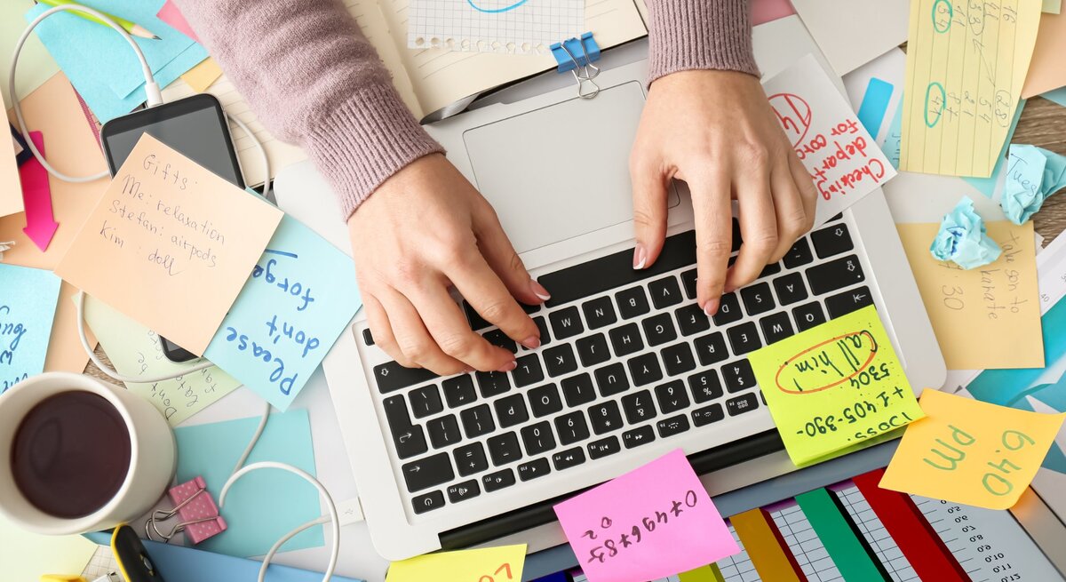 Eine Person tippt auf der Tastatur eines Laptops, umgeben von einem chaotischen Schreibtisch mit zahlreichen Haftnotizen, Dokumenten, einem Smartphone, einem Kaffeebecher und farbigen Diagrammen. Der Arbeitsplatz wirkt unorganisiert, mit Kabeln und zerknülltem Papier, was auf eine stressige oder überladene Arbeitssituation hinweist.
