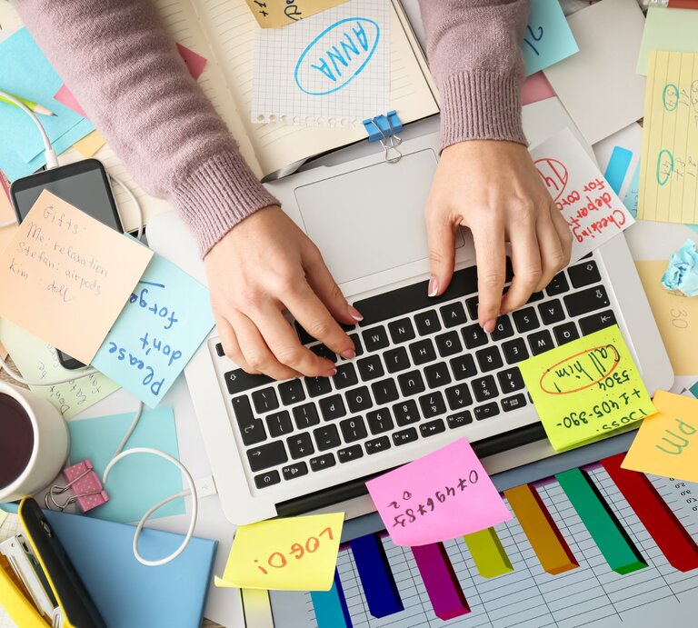 Eine Person tippt auf der Tastatur eines Laptops, umgeben von einem chaotischen Schreibtisch mit zahlreichen Haftnotizen, Dokumenten, einem Smartphone, einem Kaffeebecher und farbigen Diagrammen. Der Arbeitsplatz wirkt unorganisiert, mit Kabeln und zerknülltem Papier, was auf eine stressige oder überladene Arbeitssituation hinweist. | © Africa images via Canva.com