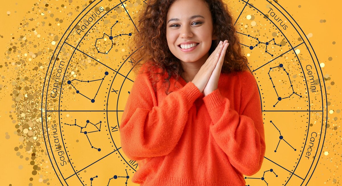 Eine junge Frau mit lockigem braunen Haar und einem orangefarbenen Pullover lächelt fröhlich und hält ihre Hände vor dem Gesicht zusammen. Hinter ihr ist ein großer astrologischer Tierkreis auf einem gelben Hintergrund zu sehen, der die Sternzeichen und ihre Konstellationen darstellt. Goldene Glitzerpunkte verstärken die visuelle Wirkung des Bildes.