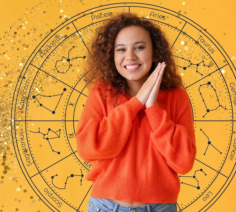 Eine junge Frau mit lockigem braunen Haar und einem orangefarbenen Pullover lächelt fröhlich und hält ihre Hände vor dem Gesicht zusammen. Hinter ihr ist ein großer astrologischer Tierkreis auf einem gelben Hintergrund zu sehen, der die Sternzeichen und ihre Konstellationen darstellt. Goldene Glitzerpunkte verstärken die visuelle Wirkung des Bildes. | © Maryna Stryzhak, pixelshot, WinWIn.artlab und Chikovnaya via Canva.com [M]