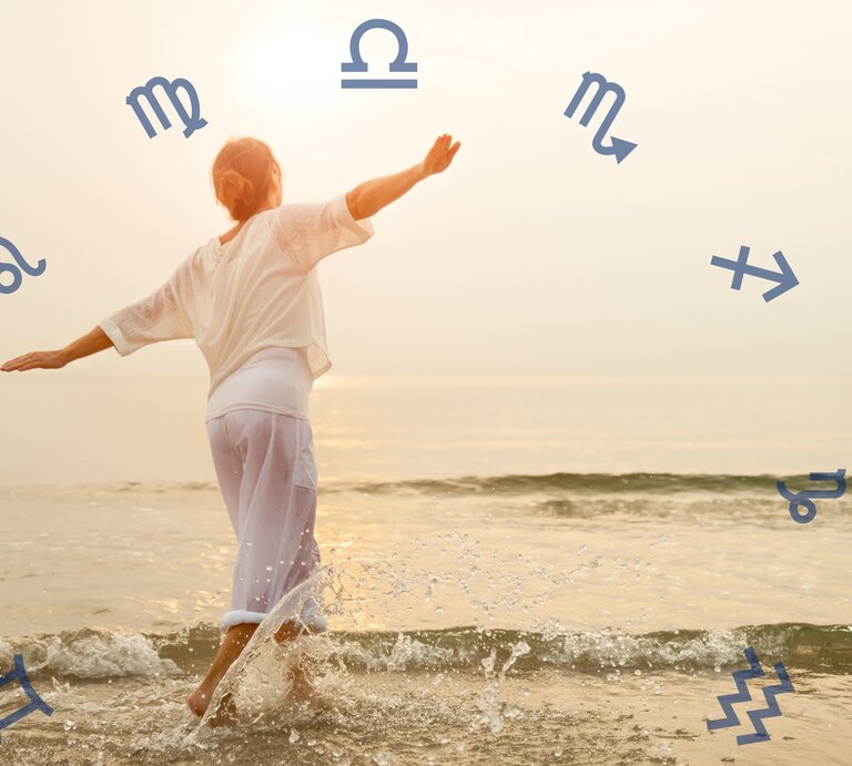 Das Bild zeigt eine Person, die an einem Strand steht und ins Wasser tritt. Sie trägt lockere weiße Kleidung und breitet die Arme aus, als würde sie die Freiheit und die Natur genießen. Um die Person herum sind verschiedene astrologische Symbole in einem Kreis angeordnet. Die Szene ist in warmes, weiches Licht getaucht, was eine friedliche und harmonische Atmosphäre erzeugt. | © Getty Images Signature/ FredFroese und Chikovnaya via Canva.com [M] 