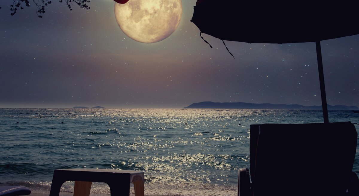 Ein ruhiger Strand, beleuchtet vom sanften Schein des Vollmonds, der majestätisch über dem Horizont aufsteigt. Der Mond wirft sein silbriges Licht auf das Meer, das sanft in kleinen Wellen an den Strand spült und dabei glitzernde Reflexionen auf der Wasseroberfläche erzeugt. Im Vordergrund stehen zwei Liegestühle und ein kleiner Tisch, die unter den Schatten von großen Sonnenschirmen verborgen sind. Der Strand ist leer und still, die Atmosphäre ruhig und verträumt, als ob die Welt für einen Moment innehält, um die Magie dieser Mondnacht zu genießen. Die entfernten Inseln am Horizont wirken wie stille Wächter dieser friedlichen Szene. Es ist ein Moment der Besinnung, des Innehaltens und des Einsseins mit der Natur.