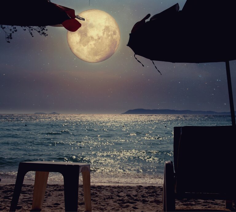 Ein ruhiger Strand, beleuchtet vom sanften Schein des Vollmonds, der majestätisch über dem Horizont aufsteigt. Der Mond wirft sein silbriges Licht auf das Meer, das sanft in kleinen Wellen an den Strand spült und dabei glitzernde Reflexionen auf der Wasseroberfläche erzeugt. Im Vordergrund stehen zwei Liegestühle und ein kleiner Tisch, die unter den Schatten von großen Sonnenschirmen verborgen sind. Der Strand ist leer und still, die Atmosphäre ruhig und verträumt, als ob die Welt für einen Moment innehält, um die Magie dieser Mondnacht zu genießen. Die entfernten Inseln am Horizont wirken wie stille Wächter dieser friedlichen Szene. Es ist ein Moment der Besinnung, des Innehaltens und des Einsseins mit der Natur. | © Getty Images/ jakkapan21 via Canva.com [M]