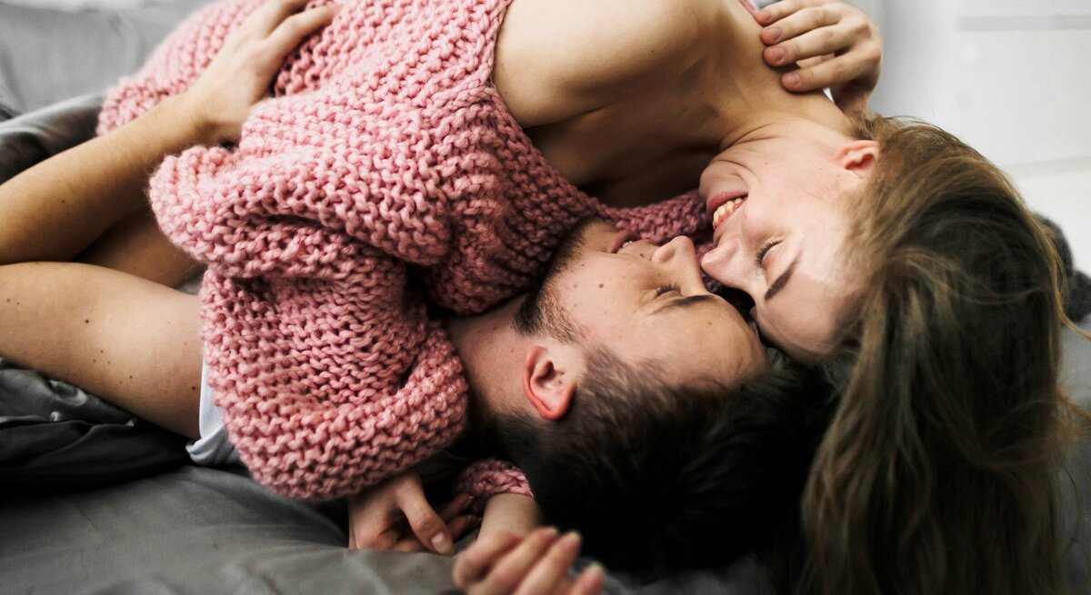 Ein verliebtes Paar liegt lachend und eng umschlungen auf einem Bett. Die Frau trägt einen grob gestrickten rosa Pullover und liegt über dem Mann, der sie liebevoll in den Armen hält. Beide wirken entspannt und glücklich, während sie sich nah sind.