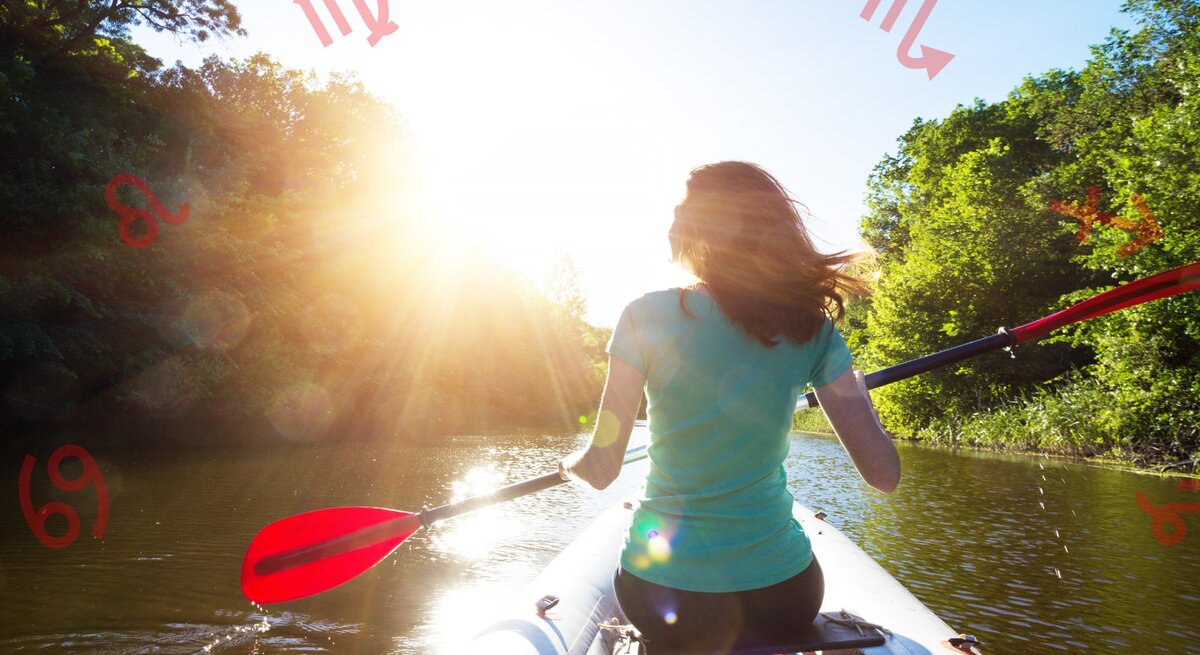 Das Bild zeigt eine Frau, die in einem Kajak auf einem ruhigen Fluss paddelt. Sie trägt ein hellblaues T-Shirt und hat mittellanges braunes Haar, das im Wind weht. Die Sonne scheint hell und strahlt durch die Bäume auf beiden Seiten des Flusses. Rote astrologische Symbole für verschiedene Sternzeichen sind über das Bild verteilt. Die Frau paddelt mit roten Paddeln und bewegt sich flussabwärts in Richtung der Sonnenstrahlen. Die Szene vermittelt ein Gefühl von Freiheit und Naturverbundenheit.