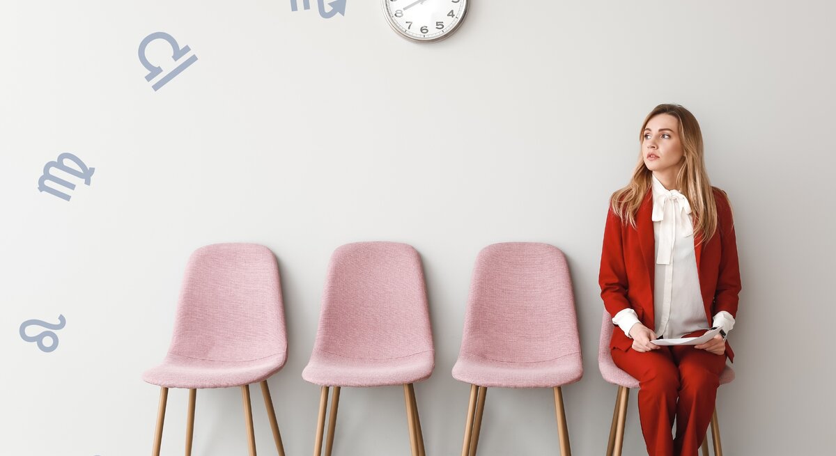 Das Bild zeigt eine junge Frau in einem eleganten, roten Hosenanzug und weißer Bluse, die auf einem rosa Stuhl sitzt und nachdenklich zur Seite blickt. Sie sitzt in einer Reihe von vier rosa Stühlen vor einer hellgrauen Wand. An der Wand hängen astrologische Symbole der Sternzeichen, die scheinbar um eine Uhr herum angeordnet sind.