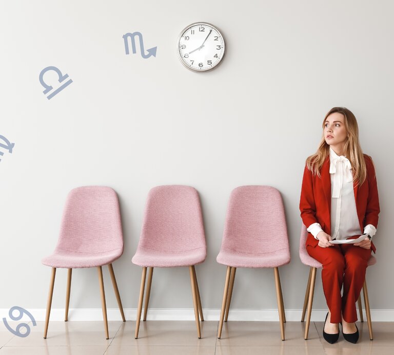 Das Bild zeigt eine junge Frau in einem eleganten, roten Hosenanzug und weißer Bluse, die auf einem rosa Stuhl sitzt und nachdenklich zur Seite blickt. Sie sitzt in einer Reihe von vier rosa Stühlen vor einer hellgrauen Wand. An der Wand hängen astrologische Symbole der Sternzeichen, die scheinbar um eine Uhr herum angeordnet sind. | © Chikovnaya und pixelshot via Canva.com [M]