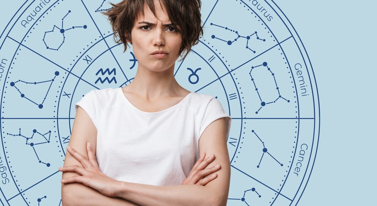 Das Bild zeigt eine junge Frau mit kurzen, braunen Haaren und einem verärgerten Gesichtsausdruck. Sie trägt ein weißes T-Shirt und hat die Arme verschränkt. Im Hintergrund ist ein astrologisches Rad mit den Sternzeichen und deren Konstellationen auf einem hellblauen Hintergrund abgebildet.