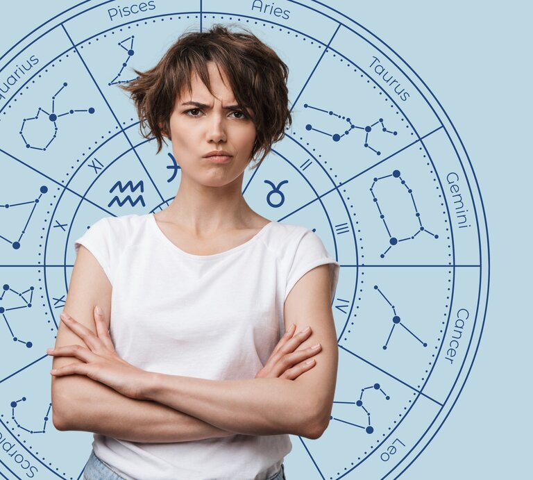 Das Bild zeigt eine junge Frau mit kurzen, braunen Haaren und einem verärgerten Gesichtsausdruck. Sie trägt ein weißes T-Shirt und hat die Arme verschränkt. Im Hintergrund ist ein astrologisches Rad mit den Sternzeichen und deren Konstellationen auf einem hellblauen Hintergrund abgebildet. | © Dean Drobot und WinWin.artlab via Canva.com [M] 