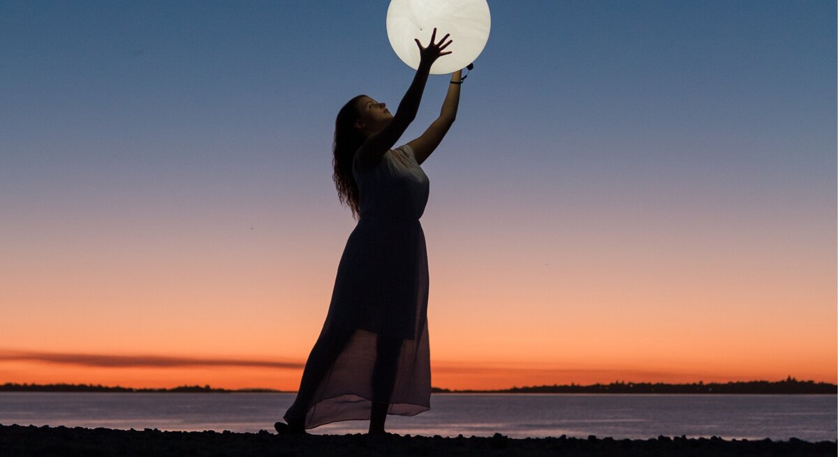 Das Bild zeigt eine Frau im langen Kleid, die am Strand steht und ihre Arme hochhält, als würde sie den großen, leuchtenden Vollmond über ihrem Kopf halten. Der Himmel im Hintergrund ist während des Sonnenuntergangs in wunderschönen Blau- und Orangetönen gefärbt, was eine ruhige und magische Atmosphäre schafft. Die Silhouette der Frau und die Reflexion des Lichts auf dem Wasser verstärken die mystische Stimmung des Bildes. Es vermittelt den Eindruck von Harmonie und Verbindung mit der Natur, passend zu einem Mondhoroskop-Thema.