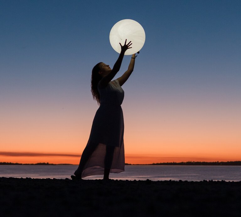 Das Bild zeigt eine Frau im langen Kleid, die am Strand steht und ihre Arme hochhält, als würde sie den großen, leuchtenden Vollmond über ihrem Kopf halten. Der Himmel im Hintergrund ist während des Sonnenuntergangs in wunderschönen Blau- und Orangetönen gefärbt, was eine ruhige und magische Atmosphäre schafft. Die Silhouette der Frau und die Reflexion des Lichts auf dem Wasser verstärken die mystische Stimmung des Bildes. Es vermittelt den Eindruck von Harmonie und Verbindung mit der Natur, passend zu einem Mondhoroskop-Thema. | © Pexels/ Ruvim