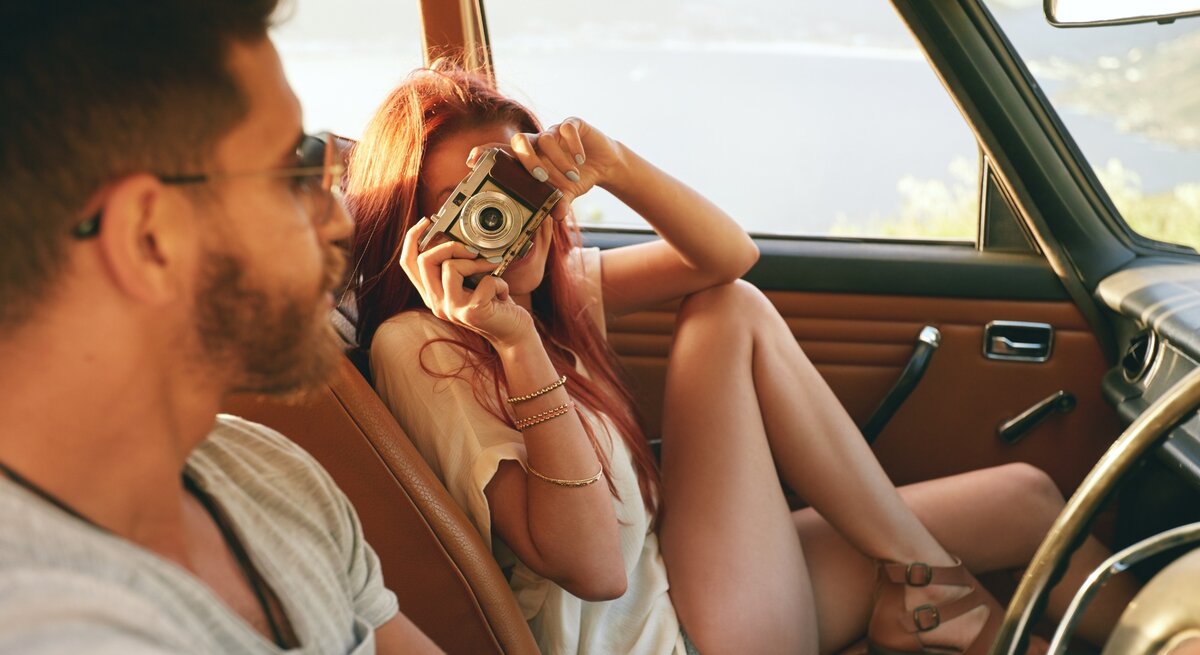 Das Bild zeigt ein Paar in einem Auto, das offensichtlich Spaß zusammen hat. Die Frau, die Shorts und ein leichtes Oberteil trägt, sitzt auf dem Beifahrersitz und hält eine Kamera vor ihr Gesicht, um ein Foto zu machen. Der Mann, der ein graues T-Shirt und eine Sonnenbrille trägt, sitzt am Steuer und schaut lächelnd zu ihr hinüber. Das Auto hat eine braune Lederausstattung und im Hintergrund ist eine malerische Landschaft mit einem See oder Meer und Hügeln zu sehen, was eine entspannte und fröhliche Stimmung vermittelt.