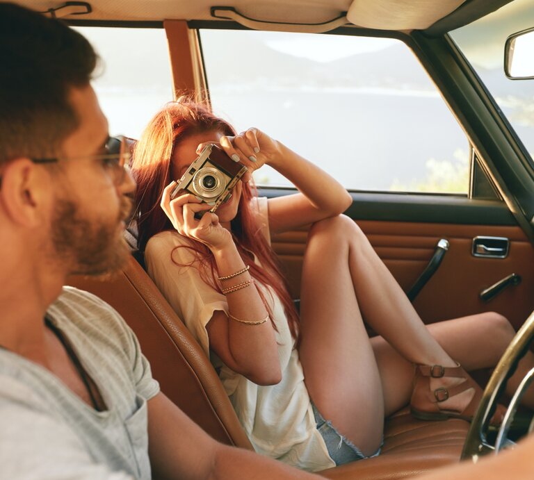 Das Bild zeigt ein Paar in einem Auto, das offensichtlich Spaß zusammen hat. Die Frau, die Shorts und ein leichtes Oberteil trägt, sitzt auf dem Beifahrersitz und hält eine Kamera vor ihr Gesicht, um ein Foto zu machen. Der Mann, der ein graues T-Shirt und eine Sonnenbrille trägt, sitzt am Steuer und schaut lächelnd zu ihr hinüber. Das Auto hat eine braune Lederausstattung und im Hintergrund ist eine malerische Landschaft mit einem See oder Meer und Hügeln zu sehen, was eine entspannte und fröhliche Stimmung vermittelt. | © Jacob Lund via Canva.com