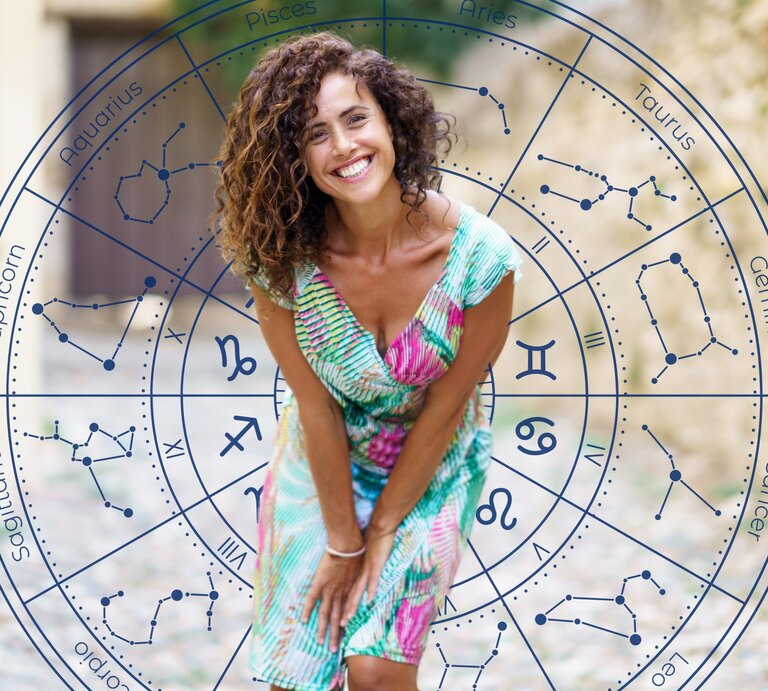 Eine lachende Frau mit lockigem Haar trägt ein buntes Sommerkleid und steht draußen auf einem gepflasterten Weg. Im Hintergrund befindet sich ein astrologischer Kreis mit den Symbolen und Konstellationen der zwölf Tierkreiszeichen, dargestellt in Blau. | © Javier Sánchez Mingorance und WinWin.artlab via Canva.com [M]