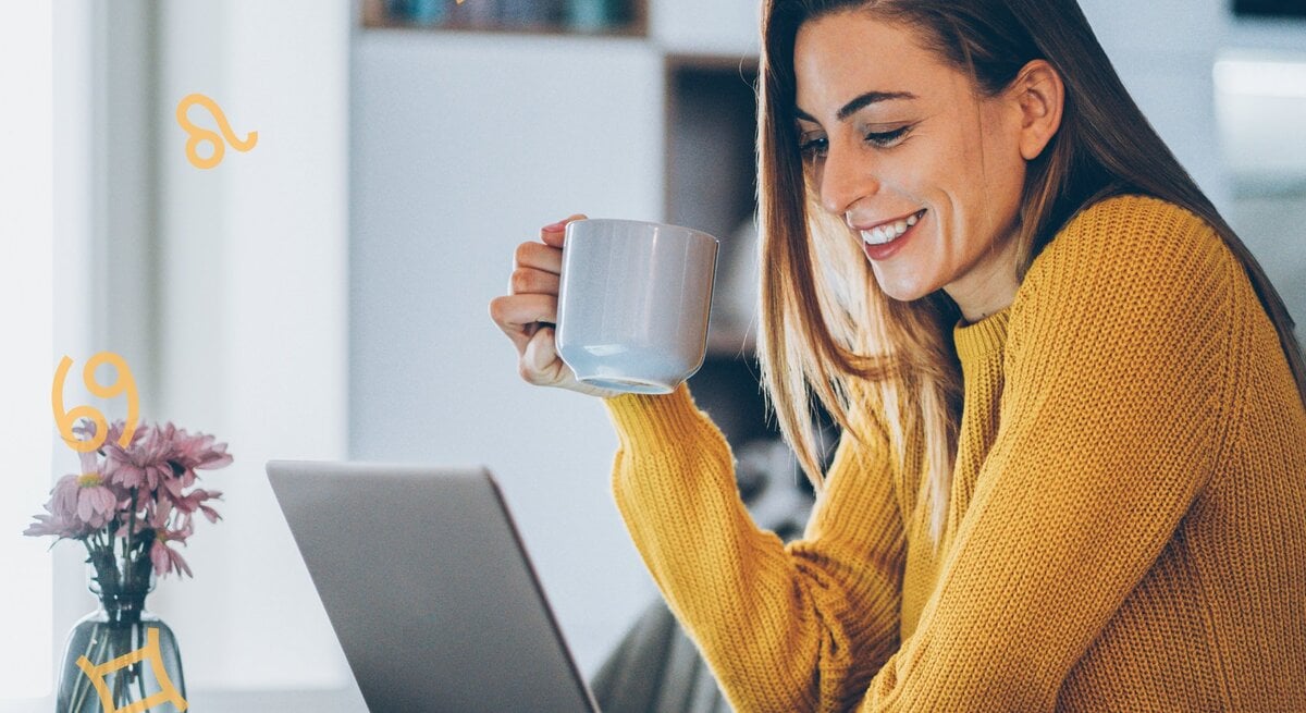 Eine Frau mit langen, braunen Haaren sitzt lächelnd an einem Tisch und schaut auf einen Laptop. Sie trägt einen gelben Pullover und hält eine Tasse in der Hand. Auf dem Tisch steht eine kleine Vase mit lila Blumen. Um sie herum schweben astrologische Symbole, die verschiedene Sternzeichen darstellen. Die Umgebung deutet auf eine gemütliche Wohnsituation hin, möglicherweise eine Küche oder ein Wohnzimmer im Hintergrund.