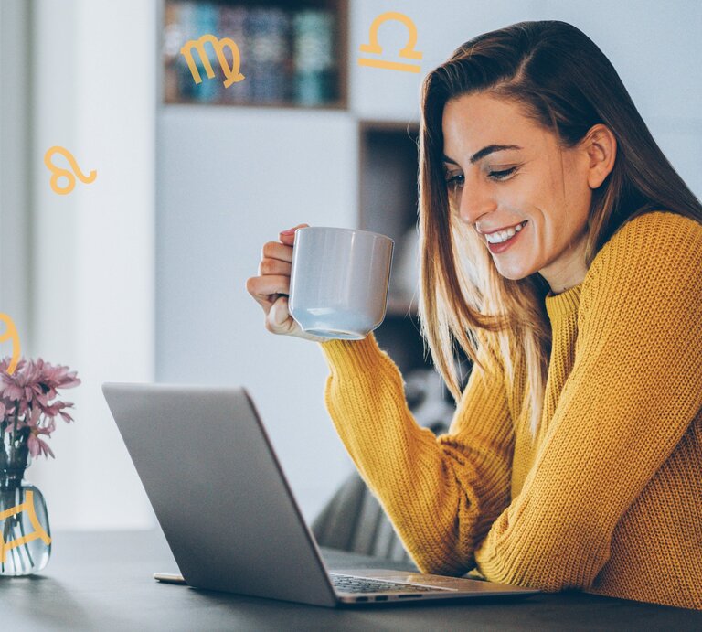 Eine Frau mit langen, braunen Haaren sitzt lächelnd an einem Tisch und schaut auf einen Laptop. Sie trägt einen gelben Pullover und hält eine Tasse in der Hand. Auf dem Tisch steht eine kleine Vase mit lila Blumen. Um sie herum schweben astrologische Symbole, die verschiedene Sternzeichen darstellen. Die Umgebung deutet auf eine gemütliche Wohnsituation hin, möglicherweise eine Küche oder ein Wohnzimmer im Hintergrund. | © Getty Images Signature/ filadendron und Chikovnaya via Canva.com [M]