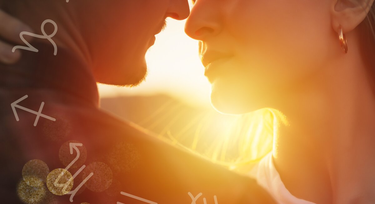 Ein Mann und eine Frau stehen sich sehr nah gegenüber, beinahe in einer Umarmung, und schauen sich intensiv in die Augen. Die Szene wird von warmem, goldenem Sonnenlicht durchflutet, das von hinten auf die beiden Personen fällt und eine romantische Atmosphäre schafft. Um sie herum sind astrologische Symbole verteilt, die auf verschiedene Sternzeichen hinweisen. Der Mann hat einen leichten Bart und trägt eine dunkle Jacke, während die Frau goldene Creolen trägt und ein weißes Oberteil.