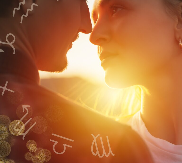 Ein Mann und eine Frau stehen sich sehr nah gegenüber, beinahe in einer Umarmung, und schauen sich intensiv in die Augen. Die Szene wird von warmem, goldenem Sonnenlicht durchflutet, das von hinten auf die beiden Personen fällt und eine romantische Atmosphäre schafft. Um sie herum sind astrologische Symbole verteilt, die auf verschiedene Sternzeichen hinweisen. Der Mann hat einen leichten Bart und trägt eine dunkle Jacke, während die Frau goldene Creolen trägt und ein weißes Oberteil. | © Adobe Stock/ eduard, Saga Design Studio und Keronyart's Images/ Be keronyart via Canva.com [M]