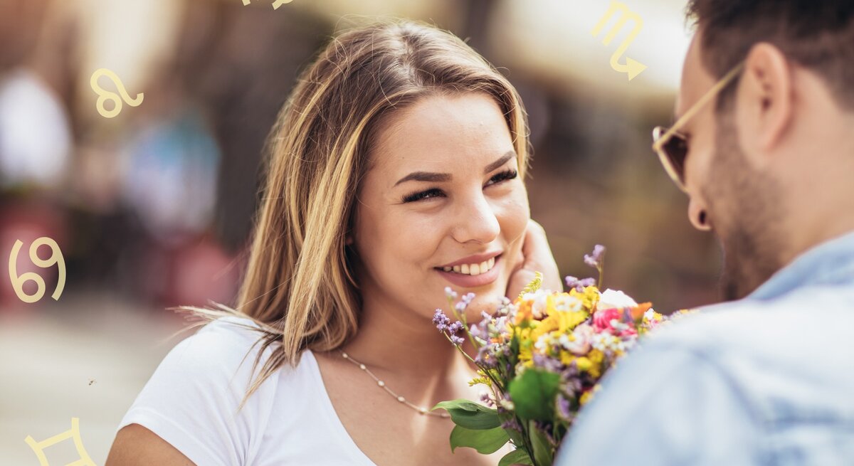 Ein Mann und eine Frau stehen sich gegenüber und lächeln sich an. Der Mann hält einen Blumenstrauß in der Hand, den er der Frau überreicht. Im Hintergrund sind verschwommene Menschen und Gebäude zu sehen. Um die Köpfe der beiden Personen sind verschiedene astrologische Symbole angeordnet, die auf die Sternzeichen hinweisen. Die Frau trägt ein weißes T-Shirt und hat blonde Haare, während der Mann eine Sonnenbrille und ein blaues Hemd trägt.