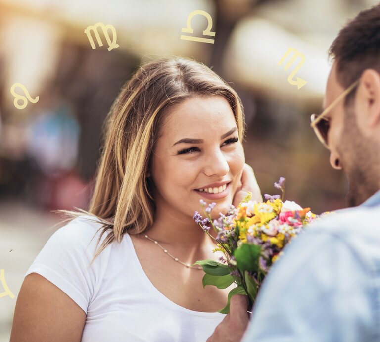Ein Mann und eine Frau stehen sich gegenüber und lächeln sich an. Der Mann hält einen Blumenstrauß in der Hand, den er der Frau überreicht. Im Hintergrund sind verschwommene Menschen und Gebäude zu sehen. Um die Köpfe der beiden Personen sind verschiedene astrologische Symbole angeordnet, die auf die Sternzeichen hinweisen. Die Frau trägt ein weißes T-Shirt und hat blonde Haare, während der Mann eine Sonnenbrille und ein blaues Hemd trägt. | © Adobe Stock/ Mediteraneo und Chikovnaya via Canva.com [M]