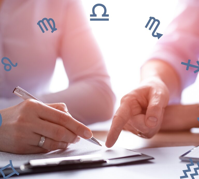 Zwei Personen in Business-Kleidung sind in ein Gespräch vertieft und arbeiten an einem Dokument. Die Hände einer Person halten einen Stift und zeigen auf das Papier, während die andere Person etwas darauf zeigt. Um das Bild herum sind verschiedene astrologische Symbole verstreut, darunter Symbole für Löwe, Jungfrau, Waage, Skorpion, Schütze, Steinbock, Wassermann und Fische. Die Szene ist in weichem Licht gehalten und hat eine professionelle Atmosphäre. | © arturmarciniecphotos und Chikovnaya via Canvas.com [M]