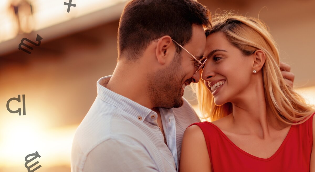 Ein lächelndes Paar steht einander nahe und genießt einen romantischen Moment im Sonnenuntergang. Der Mann trägt eine Brille und ein helles Hemd, während die Frau ein rotes Kleid trägt. Sie berühren sanft ihre Stirnen und strahlen Freude und Zuneigung aus. Im Hintergrund sind astrologische Symbole zu sehen, was eine symbolische und astrologische Bedeutung suggeriert. Die warme Beleuchtung und der unscharfe Hintergrund verstärken die intime und liebevolle Atmosphäre.