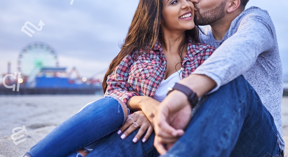 Ein junges Paar sitzt am Strand, der Mann küsst die Frau liebevoll auf die Wange. Im Hintergrund ist ein Riesenrad und eine Kirmesbeleuchtung zu sehen, was eine entspannte und romantische Atmosphäre schafft. Über dem Bild schweben verschiedene astrologische Symbole. Die Frau trägt ein kariertes Hemd und zerrissene Jeans, der Mann ein graues Langarmshirt und ebenfalls Jeans. Beide wirken glücklich und genießen den Moment.