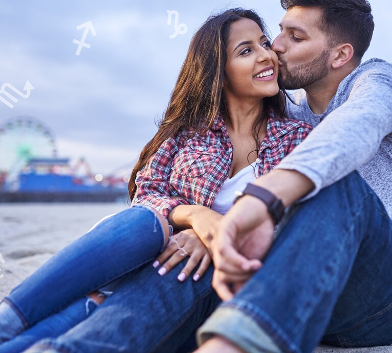 Ein junges Paar sitzt am Strand, der Mann küsst die Frau liebevoll auf die Wange. Im Hintergrund ist ein Riesenrad und eine Kirmesbeleuchtung zu sehen, was eine entspannte und romantische Atmosphäre schafft. Über dem Bild schweben verschiedene astrologische Symbole. Die Frau trägt ein kariertes Hemd und zerrissene Jeans, der Mann ein graues Langarmshirt und ebenfalls Jeans. Beide wirken glücklich und genießen den Moment. | © Joshua Resnick und Chikovnaya via Canva.com [M]