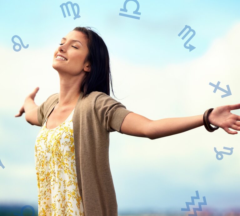 Eine lächelnde Frau steht mit ausgebreiteten Armen im Freien unter einem blauen Himmel. Sie trägt ein weißes Kleid mit gelbem Blumenmuster und eine beige Strickjacke. Um sie herum schweben verschiedene astrologische Symbole, die auf den Tierkreis hinweisen. Die Frau wirkt glücklich und frei. | © Peopleimages.com/ YuriArcus und Chikovnaya via Canva.com [M]