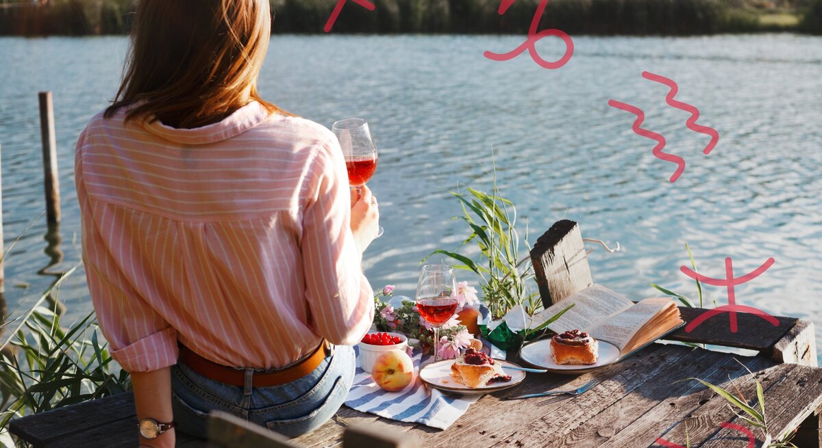 Eine Frau sitzt mit dem Rücken zur Kamera auf einem hölzernen Steg am Wasser. Sie trägt ein rosafarbenes, gestreiftes Hemd und hält ein Glas Roséwein in der Hand. Vor ihr auf dem Steg liegen ein aufgeschlagenes Buch, zwei Teller mit Kuchen, ein weiteres Glas Wein, eine Schale mit Beeren und ein Pfirsich. Im Hintergrund ist eine ruhige, ländliche Landschaft zu sehen. In der Luft sind verschiedene astrologische Symbole gezeichnet: Schütze, Steinbock, Wassermann, Fische und Widder.