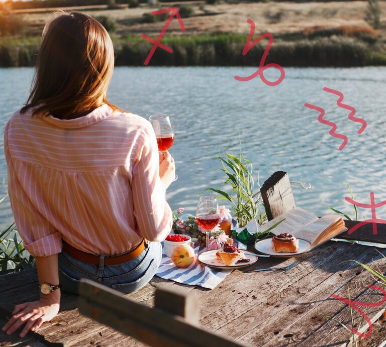 Eine Frau sitzt mit dem Rücken zur Kamera auf einem hölzernen Steg am Wasser. Sie trägt ein rosafarbenes, gestreiftes Hemd und hält ein Glas Roséwein in der Hand. Vor ihr auf dem Steg liegen ein aufgeschlagenes Buch, zwei Teller mit Kuchen, ein weiteres Glas Wein, eine Schale mit Beeren und ein Pfirsich. Im Hintergrund ist eine ruhige, ländliche Landschaft zu sehen. In der Luft sind verschiedene astrologische Symbole gezeichnet: Schütze, Steinbock, Wassermann, Fische und Widder. | © Keronyart’s Images/ Be keronyart und sergiimostovyi via Canva.com [M]