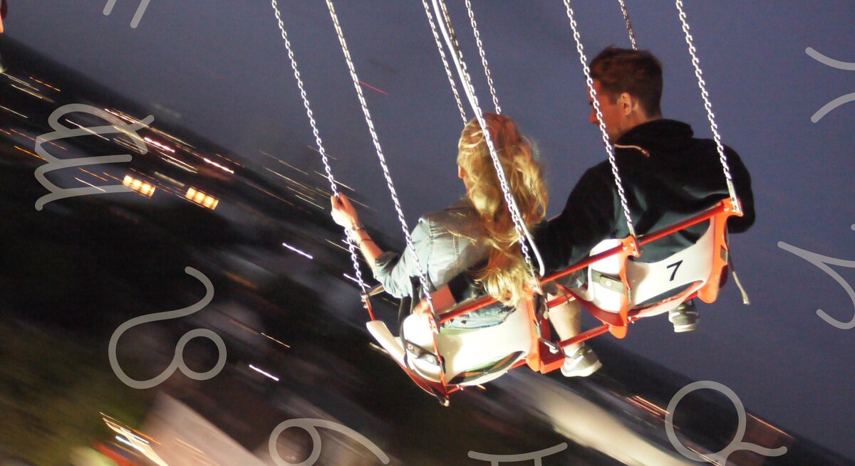 Ein Paar sitzt nebeneinander auf Schaukelsitzen eines Karussells in der Nacht. Die Frau hat lange, blonde Haare und trägt eine Jeansjacke, während der Mann dunkle Kleidung trägt. Im Hintergrund sind verschwommene Lichter einer Stadt oder eines Jahrmarkts zu sehen. Rund um das Bild sind verschiedene astrologische Symbole zu sehen: Zwillinge, Stier, Löwe, Krebs, Jungfrau und Waage.