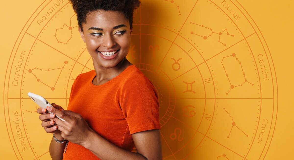 Eine Frau in einem orangefarbenen Oberteil hält ein Smartphone und lächelt. Sie steht vor einem orangefarbenen Hintergrund mit einem astrologischen Rad und Sternzeichen-Symbolen.