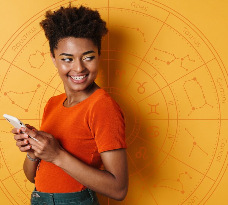 Eine Frau in einem orangefarbenen Oberteil hält ein Smartphone und lächelt. Sie steht vor einem orangefarbenen Hintergrund mit einem astrologischen Rad und Sternzeichen-Symbolen. | © Dean Drobot und WinWin.artlab via Canva.com [M]