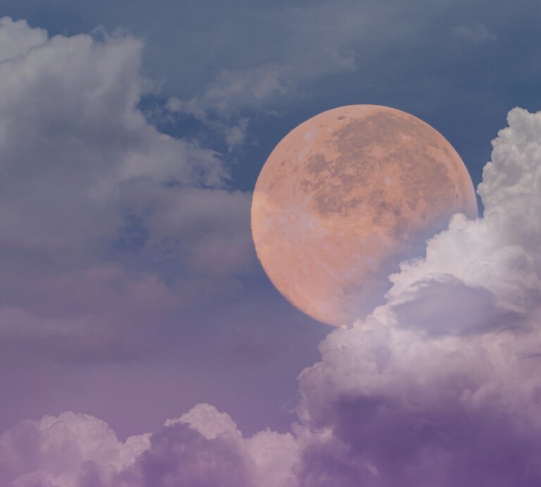 Ein großer, leuchtender Vollmond scheint durch eine teilweise bewölkte Himmelsszene. Der Himmel ist in verschiedenen Blautönen gefärbt, die von einem tiefen, dunklen Blau bis hin zu einem sanften Lila reichen. Die Wolken sind weich und flauschig, in hellen Weiß- und Lilatönen, und umrahmen den Mond, der in warmen, orangefarbenen Tönen leuchtet. Die Szenerie vermittelt eine ruhige und mystische Atmosphäre. | © Getty Images/ redonion1515