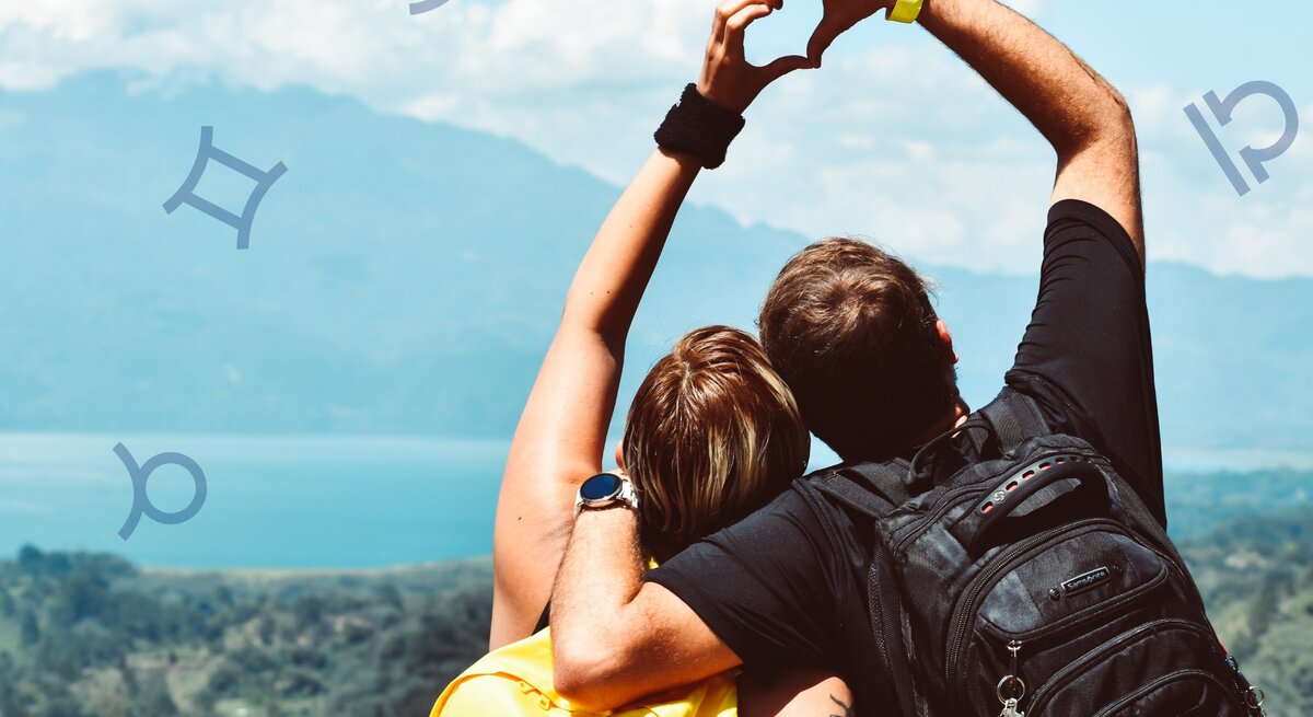 Ein Paar steht mit dem Rücken zur Kamera und blickt auf eine wunderschöne Landschaft mit Bergen und einem See. Beide tragen Rucksäcke und formen mit ihren Armen ein Herz. Um sie herum sind astrologische Symbole von verschiedenen Sternzeichen platziert.