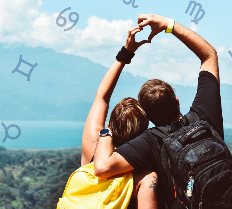 Ein Paar steht mit dem Rücken zur Kamera und blickt auf eine wunderschöne Landschaft mit Bergen und einem See. Beide tragen Rucksäcke und formen mit ihren Armen ein Herz. Um sie herum sind astrologische Symbole von verschiedenen Sternzeichen platziert. | © Pexels/ juan mendez und Chikovnaya via Canva.com [M]