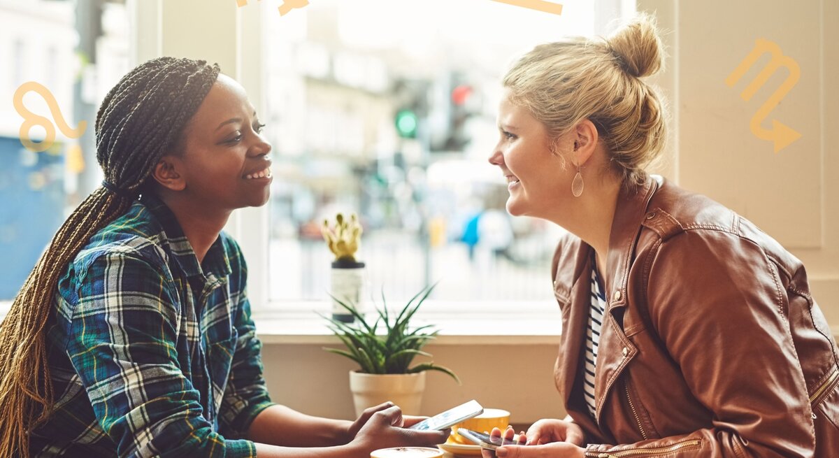 Zwei Frauen sitzen an einem Tisch in einem Café und unterhalten sich fröhlich. Die Frau links trägt ein kariertes Hemd und hat geflochtene Zöpfe, die Frau rechts trägt eine braune Lederjacke und hat blonde Haare zu einem Knoten gebunden. Im Hintergrund sind draußen unscharf Gebäude und Menschen zu sehen. Um sie herum sind astrologische Symbole von verschiedenen Sternzeichen platziert.
