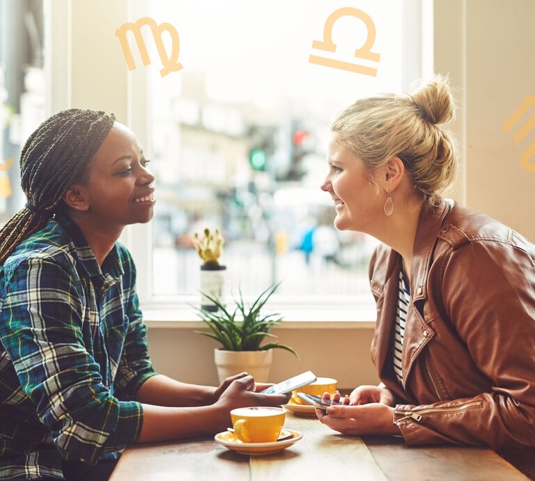 Zwei Frauen sitzen an einem Tisch in einem Café und unterhalten sich fröhlich. Die Frau links trägt ein kariertes Hemd und hat geflochtene Zöpfe, die Frau rechts trägt eine braune Lederjacke und hat blonde Haare zu einem Knoten gebunden. Im Hintergrund sind draußen unscharf Gebäude und Menschen zu sehen. Um sie herum sind astrologische Symbole von verschiedenen Sternzeichen platziert. | © Getty Images Pro/ Charday Penn und Chikovnaya via Canva.com [M]