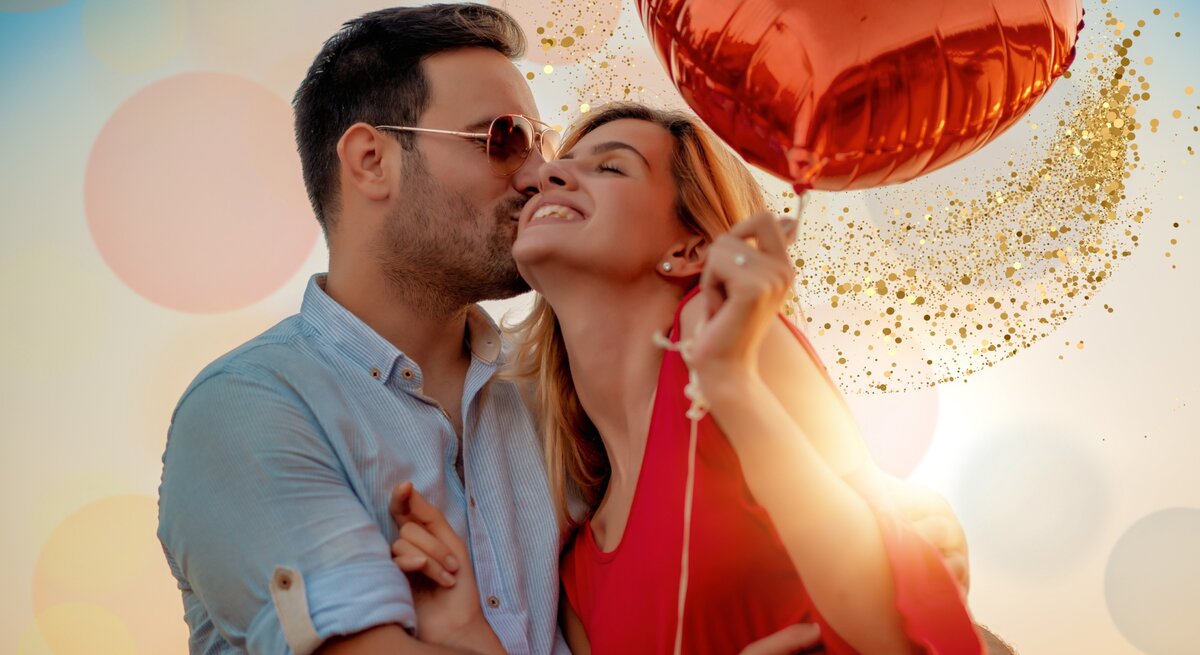 Auf dem Bild ist ein glückliches Paar zu sehen, das sich liebevoll umarmt und küsst. Der Mann trägt ein hellblaues Hemd und eine Sonnenbrille, während die Frau ein leuchtend rotes Kleid trägt und ebenfalls eine Sonnenbrille hat. Sie hält einen herzförmigen, roten Luftballon. Im Hintergrund sind verschwommene Lichtkreise und eine Glitzerwolke, die den feierlichen und romantischen Anlass unterstreicht. Das Bild strahlt Wärme und Zuneigung aus, verstärkt durch das weiche, warme Licht der Umgebung.