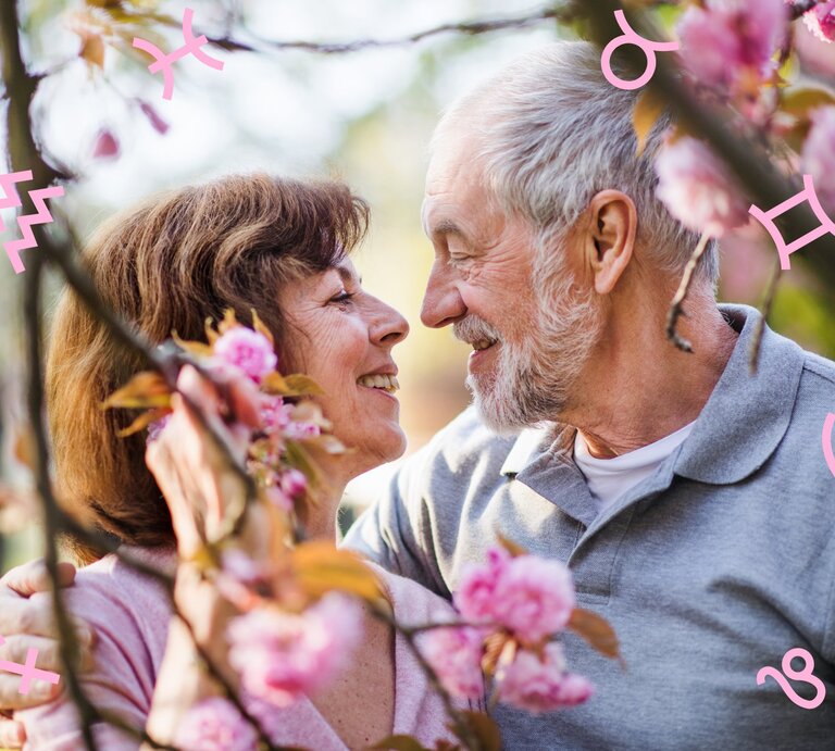 Ein älteres Paar, das liebevoll und lächelnd Augenkontakt hält. Die Frau hat braunes Haar und trägt ein rosa Oberteil, während der Mann graues Haar und einen grauen Pullover hat. Sie umarmen sich inmitten der Blüten eines Kirschblütenbaums. Grafische Symbole der Tierkreiszeichen sind über das Bild verteilt, was auf einen astrologischen oder romantischen Kontext hindeutet. | © Halfpoint und Chikovnaya via Canva.com [M]