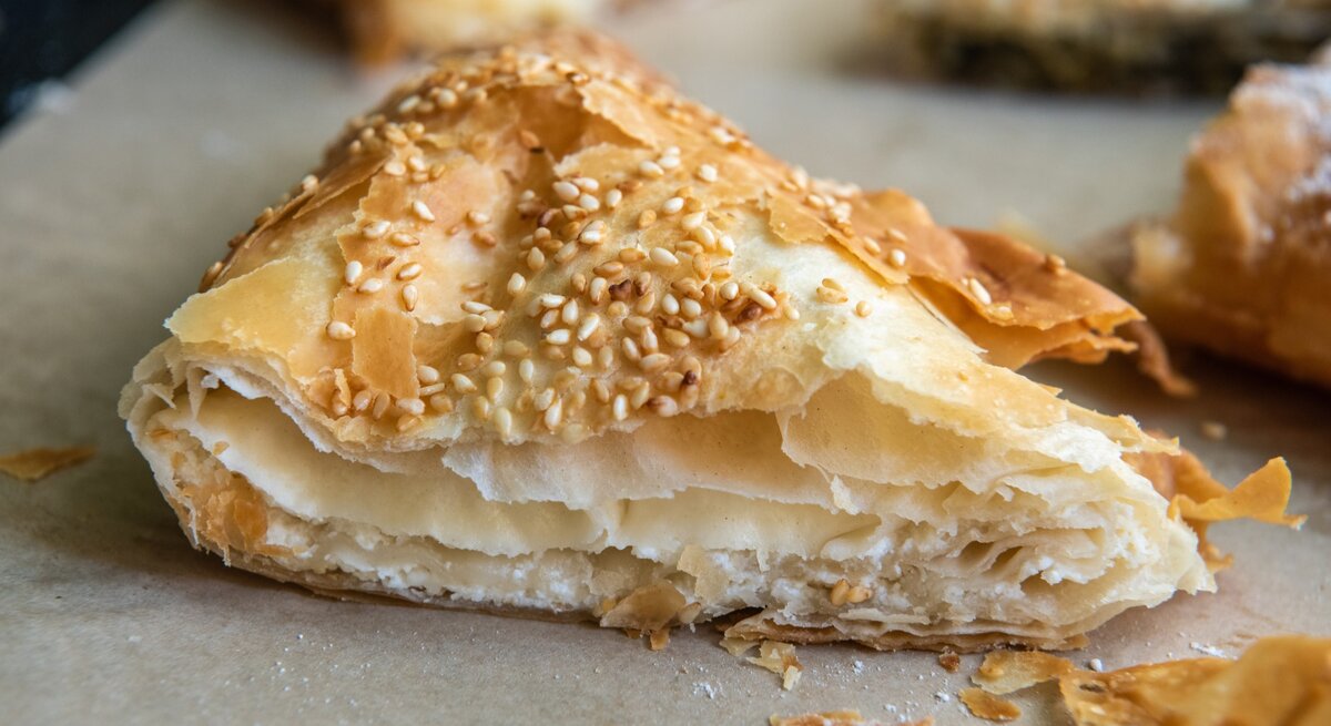Ein Stück Tiropita auf einem Blech mit Backpapier. Alles nah und von vorne fotografiert.