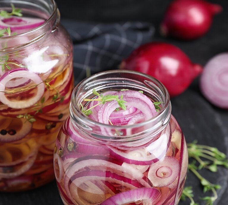 Zwei Weckgläser mit eingelegten, roten Zwiebelringen auf einem dunklen Tisch mit frischen Kräutern. | © Shutterstock/ New Africa